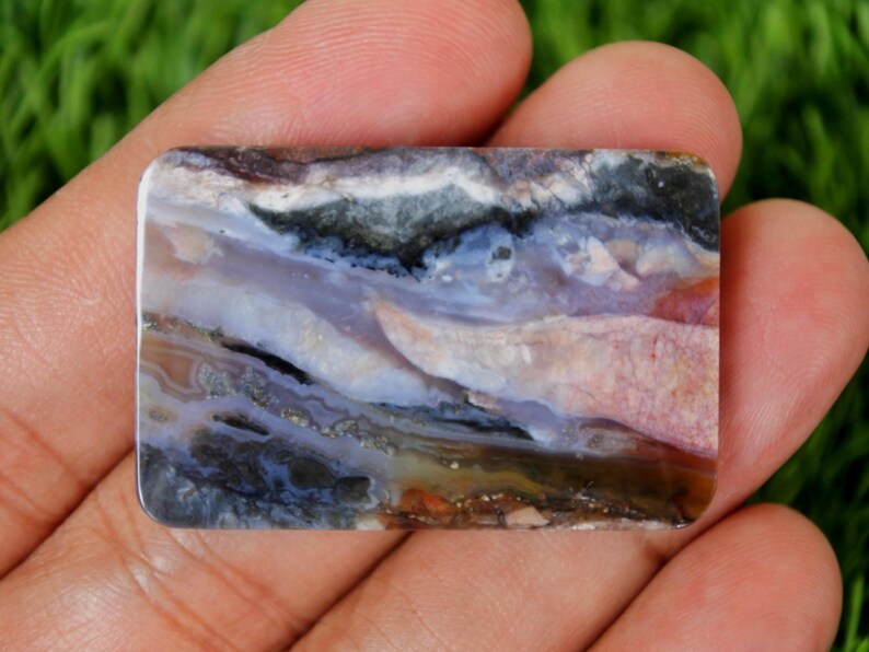 Pode incluir: Uma pedra preciosa retangular com uma superf&iacute;cie polida. A pedra apresenta uma mistura de cores, incluindo branco, preto, azul, roxo e castanho. O padr&atilde;o assemelha-se a uma paisagem com texturas em camadas e forma&ccedil;&otilde;es naturais. A pedra preciosa &eacute; segurada em uma m&atilde;o.