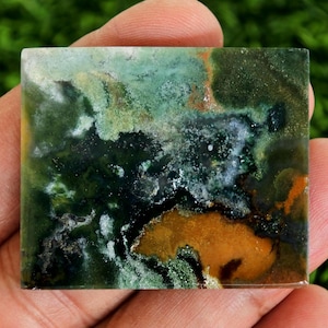 May include: A square stone featuring a blend of green, white, and orange colours. The surface displays a marbled effect, with a mix of dark and light shades. The stone has a natural, earthy appearance, ideal for jewellery or decorative use. The stone is measured in centimetres.