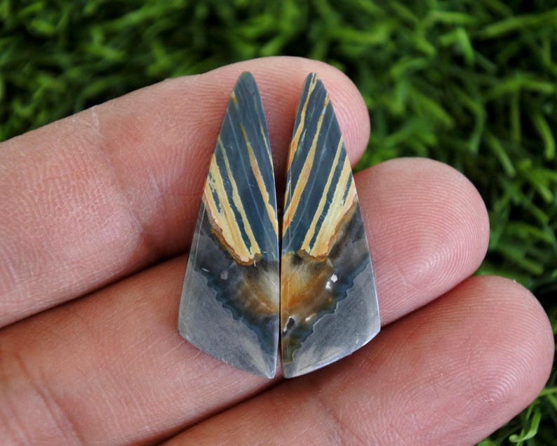 Pode incluir: Duas pedras preciosas triangulares polidas com um padr&atilde;o marcante. As pedras apresentam um gradiente de cores, incluindo azul, bege e cinza. O design assemelha-se a uma asa ou pena estilizada, com linhas paralelas.