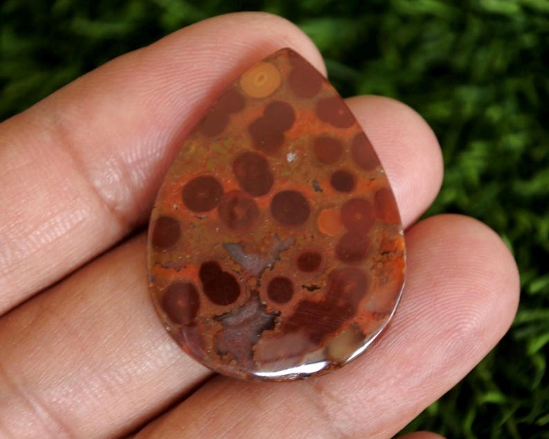 May include: A polished, teardrop-shaped cabochon of orbicular jasper featuring a smooth, glossy surface. The stone displays a rich colour palette of deep reddish-brown, burnt orange, and tan, characterised by distinct circular patterns and inclusions.