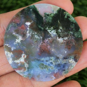 May include: A circular gemstone with a polished surface, displaying a mix of colours. The stone has green, blue, red, and white tones, forming a marbled design. The gemstone is held in a hand, highlighting its natural beauty and intricate details.