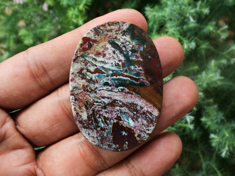 Puede incluir: Una gema ovalada con un patr&oacute;n de colores &uacute;nico. La piedra presenta una mezcla de tonos rojos, marrones, blancos y turquesas, creando un efecto veteado. La superficie pulida de la piedra refleja la luz, resaltando su belleza natural. Ideal para la creaci&oacute;n de joyas.