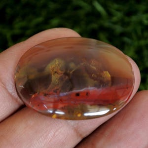 May include: An oval-shaped, polished cabochon gemstone featuring a translucent, warm amber-toned surface with internal inclusions. The stone displays distinct layers of deep red, golden yellow, and brown hues, creating a complex, organic pattern.