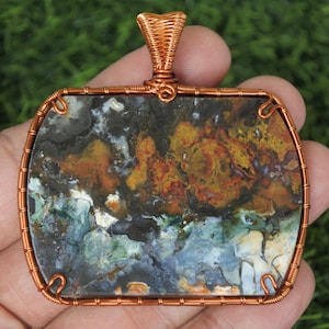 May include: A rectangular pendant with a copper wire frame and bail. The stone within the frame displays a mix of colours, including brown, yellow, blue, and white. The pendant is held in a hand against a green background.