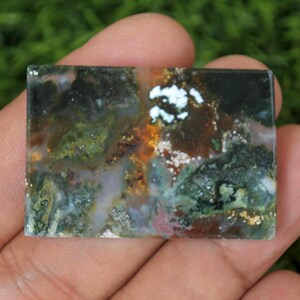 May include: A rectangular gemstone with a distinctive colour pattern. The stone displays a blend of green, brown, white, and orange tones, forming a natural, abstract design. The polished surface reflects light, accentuating the intricate details.