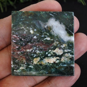 May include: Square-shaped gemstone with a natural pattern of green, white, and maroon. The stone has a polished surface and is held in a hand. The gemstone's design resembles a landscape.