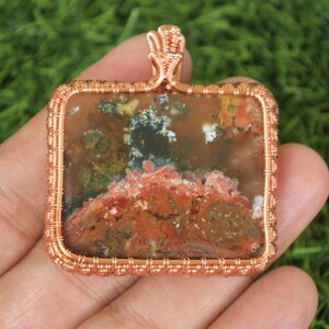 Pode incluir: Um pingente quadrado com uma moldura de arame de cobre. O pingente apresenta uma pedra natural com uma mistura de cores, incluindo vermelho, marrom, verde e branco. O arame é intrincadamente tecido ao redor da pedra, criando uma borda e uma argola decorativas.