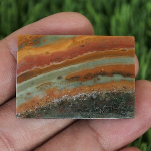 Może przedstawiać: Gładki, wypolerowany, prostokątny płat agatu wstęgowego z mieszanką zielonych, brązowych, pomarańczowych i żółtych kolorów. Agat ma warstwowy, prążkowany wzór.