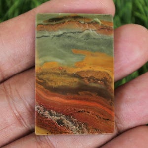 May include: A rectangular gemstone with a layered pattern of colours. The stone features bands of green, orange, brown, and red, creating a landscape-like appearance. The stone is held in a hand, showcasing its size and texture.