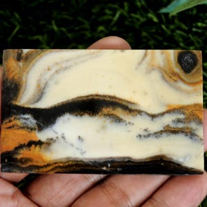 May include: A rectangular polished stone slab featuring a smooth surface with swirling patterns of cream, black, and golden-brown colors. The stone displays organic, layered bands and distinct mineral inclusions that create a natural landscape effect.