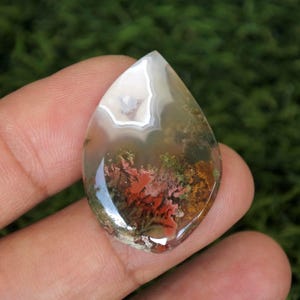 May include: A teardrop-shaped agate cabochon with a white, green, and orange dendritic pattern. The agate is translucent and shows a variety of colors and patterns.