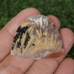 Può includere: Una gemma lucida, semicircolare, con una base traslucida. La pietra presenta intricati motivi neri, beige e bianchi, che ricordano un paesaggio. Un'area marrone-rossastra è visibile vicino al bordo inferiore. La pietra è tenuta in una mano.