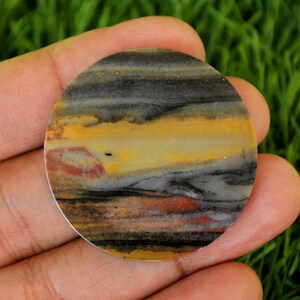 May include: A round, polished stone with a layered pattern of black, gray, yellow, and reddish-brown hues. The stone is held in a hand against a backdrop of green grass. The stone's diameter is approximately 2 inches.
