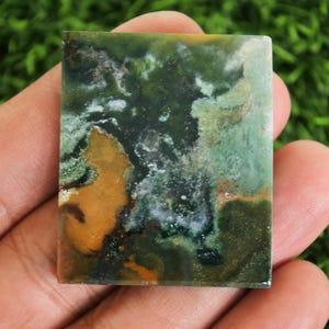 Peut inclure: Une pierre pr&eacute;cieuse rectangulaire polie. La pierre pr&eacute;sente un m&eacute;lange de teintes vertes, brunes et blanches, cr&eacute;ant un motif abstrait naturel. La pierre est tenue dans une main sur un fond de feuillage vert. Id&eacute;al pour la joaillerie.