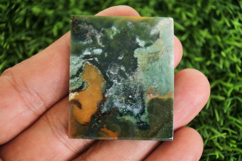 Peut inclure: Une pierre pr&eacute;cieuse rectangulaire polie. La pierre pr&eacute;sente un m&eacute;lange de teintes vertes, brunes et blanches, cr&eacute;ant un motif abstrait naturel. La pierre est tenue dans une main sur un fond de feuillage vert. Id&eacute;al pour la joaillerie.