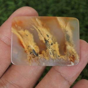 Puede incluir: Una gema rectangular con esquinas redondeadas, que muestra un patrón natural de tonos amarillos, naranjas y negros. La piedra se sostiene en una mano, con un fondo verde borroso.