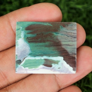 Puede incluir: Una piedra preciosa cuadrada con un patrón único de tonos verdes, marrones y blancos. La piedra tiene una apariencia veteada, con colores arremolinados y una superficie pulida. La piedra preciosa se sostiene en una mano sobre un fondo verde borroso.