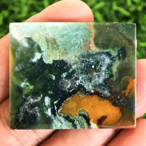 Peut inclure: Pierre pr&eacute;cieuse carr&eacute;e avec un motif unique de teintes vertes, brunes et orang&eacute;es. La pierre a une surface polie et un aspect naturel et terreux. Les couleurs se m&eacute;langent, cr&eacute;ant un design abstrait.