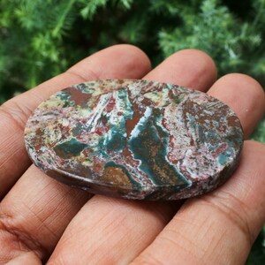 Puede incluir: Una gema ovalada con un patr&oacute;n &uacute;nico de colores rojo, verde, marr&oacute;n y blanco. La superficie pulida de la piedra muestra una est&eacute;tica natural y terrosa. La gema se sostiene en una mano, mostrando su tama&ntilde;o y textura.