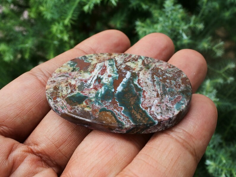 Puede incluir: Una gema ovalada con un patr&oacute;n &uacute;nico de colores rojo, verde, marr&oacute;n y blanco. La superficie pulida de la piedra muestra una est&eacute;tica natural y terrosa. La gema se sostiene en una mano, mostrando su tama&ntilde;o y textura.