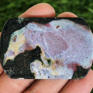 May include: A rectangular stone with rounded corners, featuring a mix of colors. The stone has a black border, with a central area of purple, white, and green hues. The stone is held in a hand.