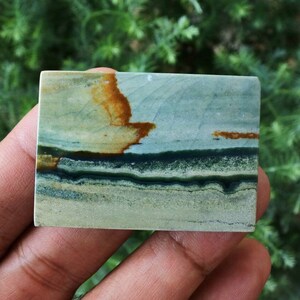 May include: A rectangular stone with a layered pattern of green, brown, and tan hues. The stone's surface has a smooth texture, and the color variations create a natural, earthy aesthetic. The stone is held in a hand.