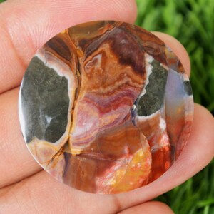 May include: A polished, round gemstone with a swirling pattern of colors. The stone features shades of brown, orange, red, purple, and green. The gemstone is held in a hand, with a green background visible.