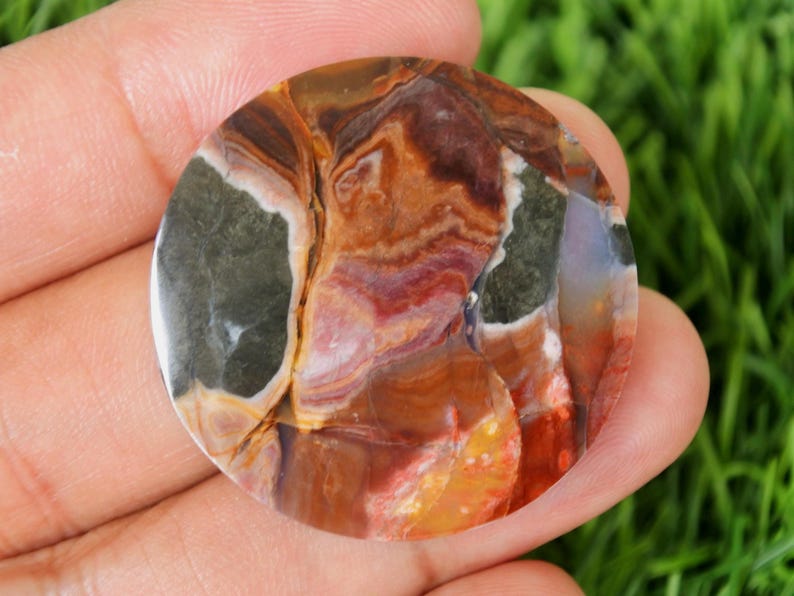 May include: A polished, round gemstone with a swirling pattern of colors. The stone features shades of brown, orange, red, purple, and green. The gemstone is held in a hand, with a green background visible.
