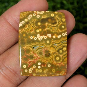 Puede incluir: Una gema rectangular con un patrón único de diseños circulares verdes oliva, amarillos y blancos. La piedra tiene una superficie pulida y se sostiene en una mano, mostrando su belleza natural. El diseño de la piedra incluye acentos rojos y marrones.