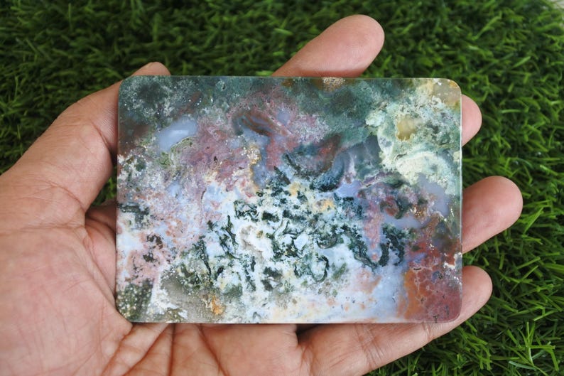 May include: A rectangular slab of polished moss agate featuring a complex pattern of deep forest green, soft lavender, and earthy terracotta inclusions. The stone displays a smooth, flat surface with translucent areas and intricate mineral dendrites.