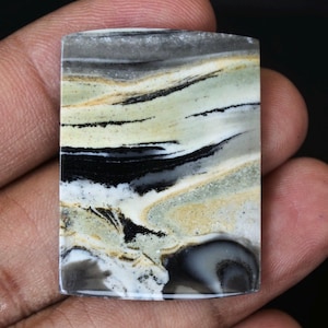 Puede incluir: Una piedra preciosa rectangular con una superficie pulida. La piedra presenta un patrón arremolinado de colores gris, negro, blanco y beige. La piedra se sostiene en una mano, mostrando su tamaño y forma. La estética general es natural y terrosa.