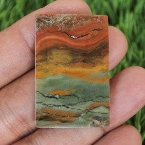 Puede incluir: Una piedra rectangular con un patrón en capas de colores. La piedra presenta bandas de rojo, naranja, amarillo, marrón y verde. La piedra se sostiene en una mano sobre un fondo de hierba verde.