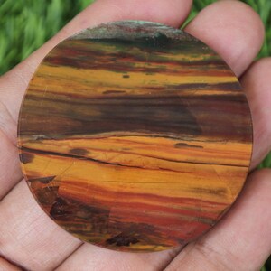 May include: A circular, polished stone displaying a natural, layered pattern in warm tones. The stone has bands of orange, brown, and red, creating a unique, earthy aesthetic. The stone is held in a hand, showing its size and texture.