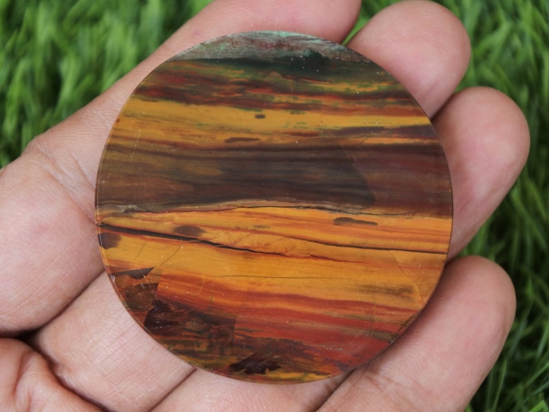 May include: A circular, polished stone displaying a natural, layered pattern in warm tones. The stone has bands of orange, brown, and red, creating a unique, earthy aesthetic. The stone is held in a hand, showing its size and texture.