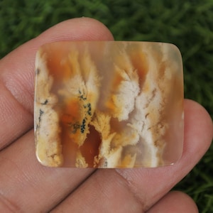 Puede incluir: Una piedra preciosa de forma cuadrada con un patrón único de inclusiones naranjas, blancas y negras. La superficie pulida de la piedra muestra un diseño natural y abstracto, que se asemeja a un paisaje o formación de nubes. La piedra se sostiene en una mano.