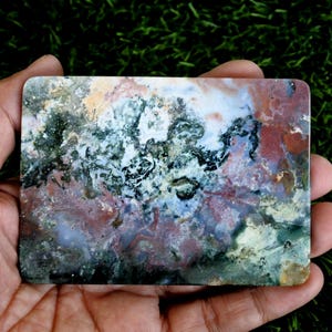 May include: A rectangular polished slab of moss agate featuring a complex pattern of deep forest green, earthy terracotta red, creamy white, and translucent gray inclusions. The stone has smooth, rounded corners and a flat, reflective surface.