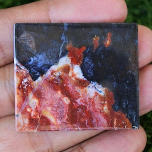 Peut inclure: Une pierre précieuse carrée et polie, présentant un motif saisissant de teintes rouges, noires et bleues. La surface de la pierre affiche un mélange de couleurs, les zones rouges et blanches contrastant avec les nuances plus sombres. La pierre est tenue dans une main.