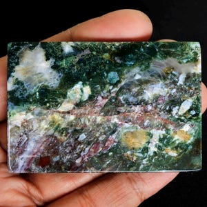 May include: A rectangular, polished gemstone with a vibrant mix of colors. The stone features a base of deep green, interspersed with white, red, and yellow hues. The intricate patterns and textures create a unique, natural aesthetic. The gemstone is held in a hand.