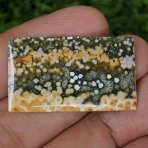 Puede incluir: Primer plano de un cabujón de jaspe pulido, rectangular, verde, blanco y amarillo. La piedra tiene un patrón moteado con manchas blancas y amarillas sobre un fondo verde.