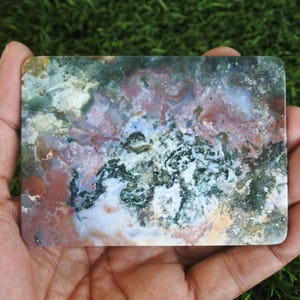 May include: A rectangular slab of polished moss agate featuring a complex, multi-coloured pattern of deep forest green, earthy terracotta red, creamy white, and translucent grey inclusions, showcasing natural mineral formations and textures.