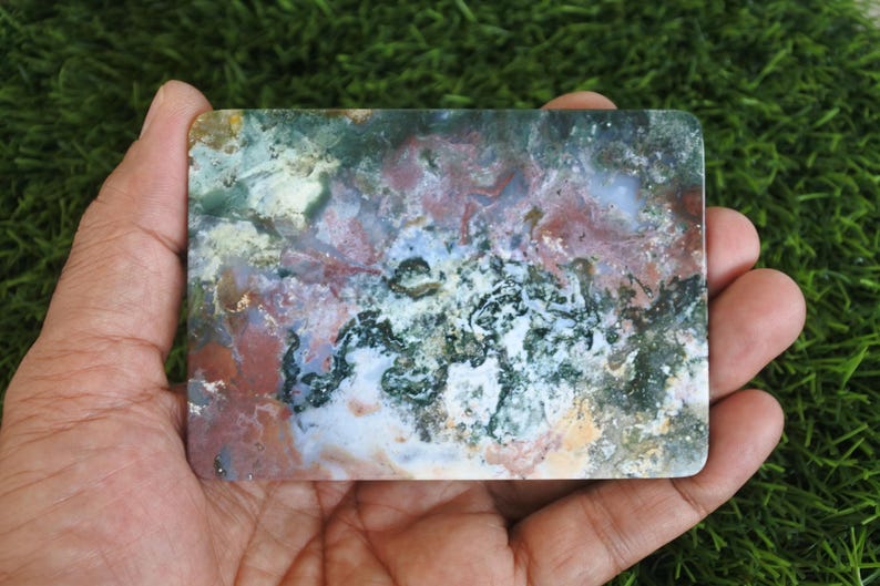 May include: A rectangular slab of polished moss agate featuring a complex, multi-coloured pattern of deep forest green, earthy terracotta red, creamy white, and translucent grey inclusions, showcasing natural mineral formations and textures.