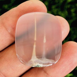 Op de afbeelding: Een doorschijnende, lichtroze edelsteen met een afgeronde rechthoekige vorm. De steen heeft een verticale, lichtgekleurde insluitsel die door het midden loopt en een vleugje groen aan de basis. De achtergrond is een vage groene kleur.