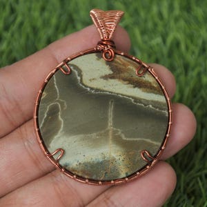 Może przedstawiać: Okrągła zawieszka z naturalnym kamieniem w środku, z odcieniami zieleni, brązu i bieli. Kamień jest osadzony w miedzianej ramie i krawędzi, tworząc ręcznie robioną biżuterię. Zawieszka jest prezentowana na zielonym tle.