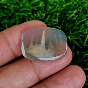 Könnte beinhalten: Ein durchscheinender weißer ovaler Cabochon-Edelstein mit einem wolkigen, weißen und grauen Muster. Der Stein ist glatt und poliert.