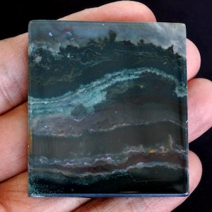 May include: A square, polished stone displaying a layered pattern of dark green, teal, and brown tones. The stone has a glossy finish and appears to be a natural mineral specimen. The colours blend, creating a unique, abstract design.