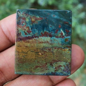 May include: A rectangular gemstone with a unique pattern of colors. The stone features a blend of teal, brown, and gold hues, with reddish-brown accents. The surface has a textured appearance, showcasing natural variations. This gemstone is ideal for jewelry making.