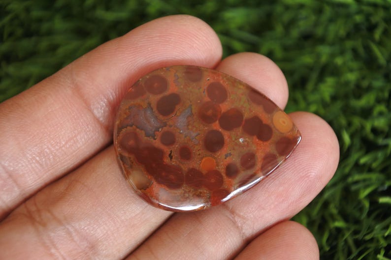 May include: A polished, teardrop-shaped orbicular jasper gemstone featuring a smooth, glossy surface with a distinct pattern of dark brown and reddish-brown circular inclusions set against a warm, earthy, terracotta-coloured background.