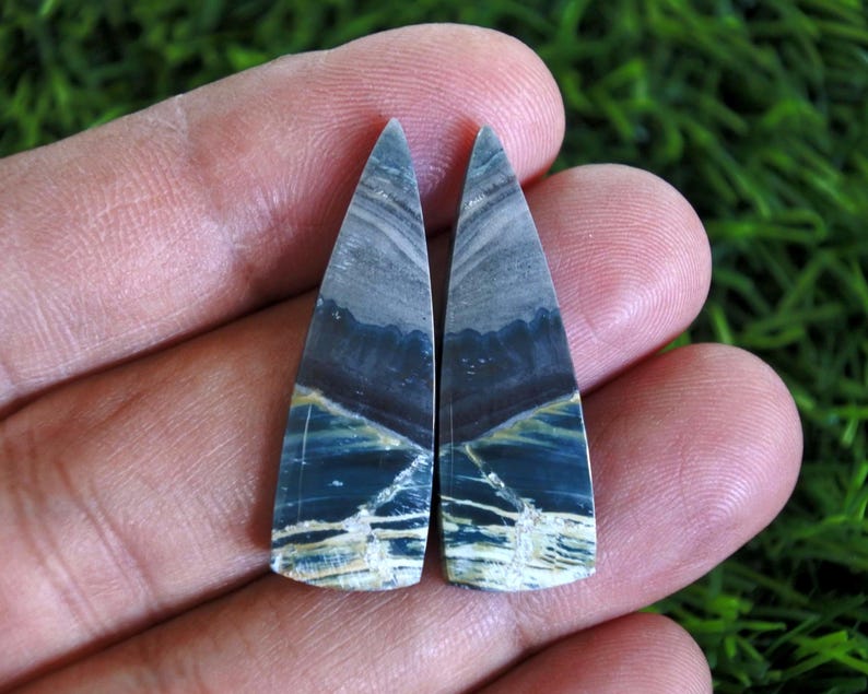 Może przedstawiać: Dwa polerowane wisiorki z kamieni szlachetnych o tr&oacute;jkątnym kształcie. Kamienie mają mieszankę kolor&oacute;w niebieskiego, szarego i kremowego z unikalnym, warstwowym wzorem. Wisiorki są trzymane w dłoni na zielonym tle.