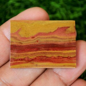 May include: A rectangular stone with a layered pattern of yellow, red, and orange hues. The stone is held in a hand, showcasing its natural colours and textures. The stone's design resembles a landscape.