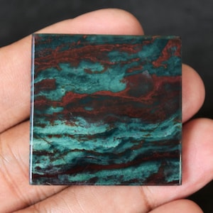 May include: Square-shaped gemstone with a polished surface. The stone features a swirling pattern of teal and maroon colors. The gemstone is held in a hand, showcasing its size and texture. Ideal for jewelry making or collecting.